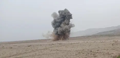 A column of dark smoke rises from a controlled explosion on flat, empty ground.
