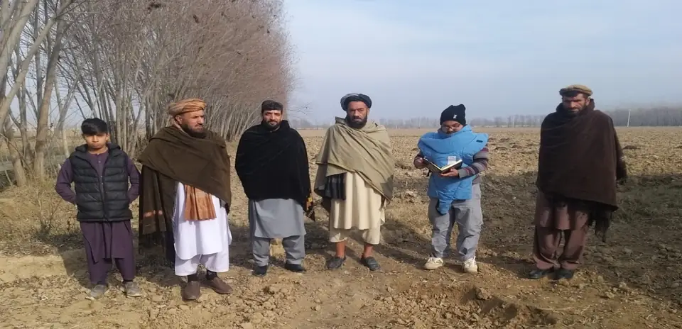 MCPA team members, one in a protective vest, stand with village elders on open ground under bare trees.