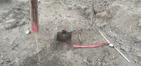 An improvised device exposed in a small excavation, with a red-and-white measuring pole and marker stakes around it.