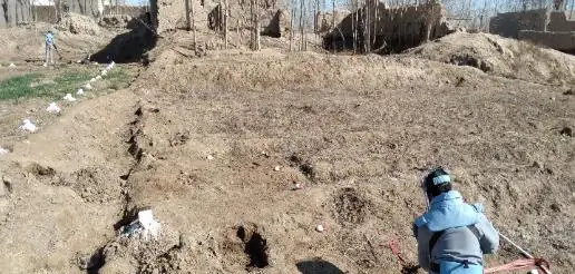 A deminer in protective equipment kneels to excavate carefully in rough ground beside a village wall.