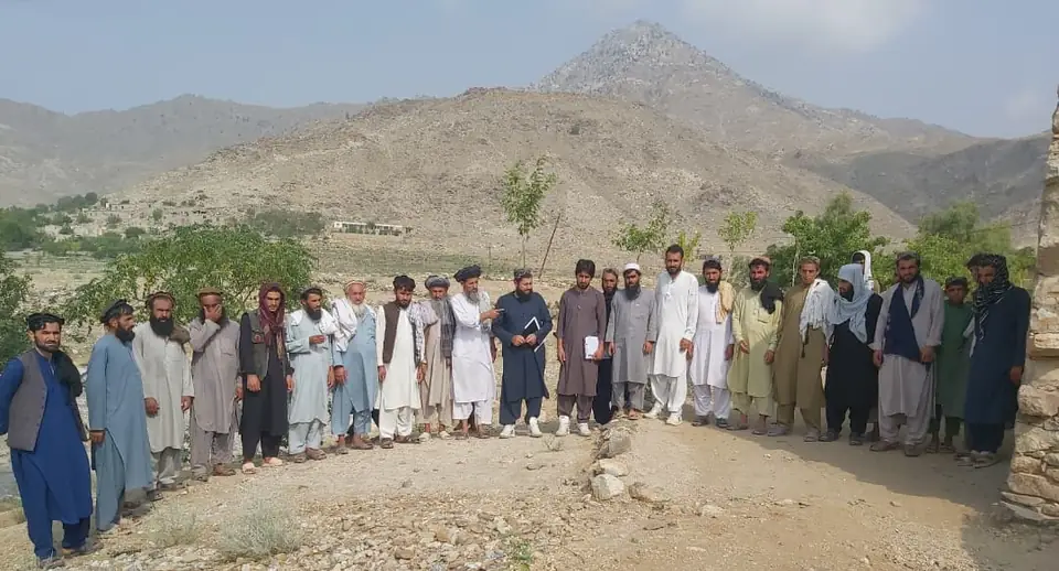 A large group of men and boys from Badpash Bar Kala stand together in a line on open ground below a mountain.