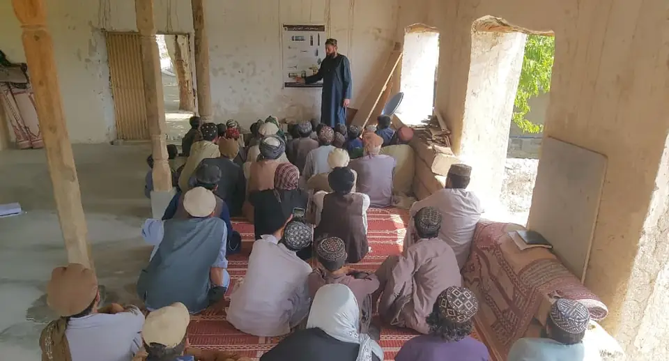 Men and boys sit on carpets in a mosque's covered courtyard, facing an MCPA educator who stands beside a poster of explosive items.