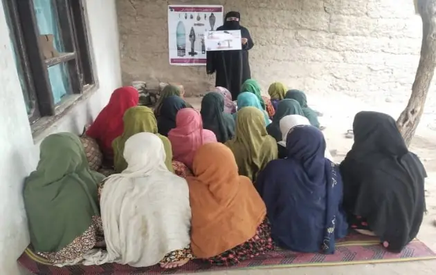 Seen from behind, a group of women in headscarves sit on the floor of a courtyard as a woman educator shows posters of explosive items.