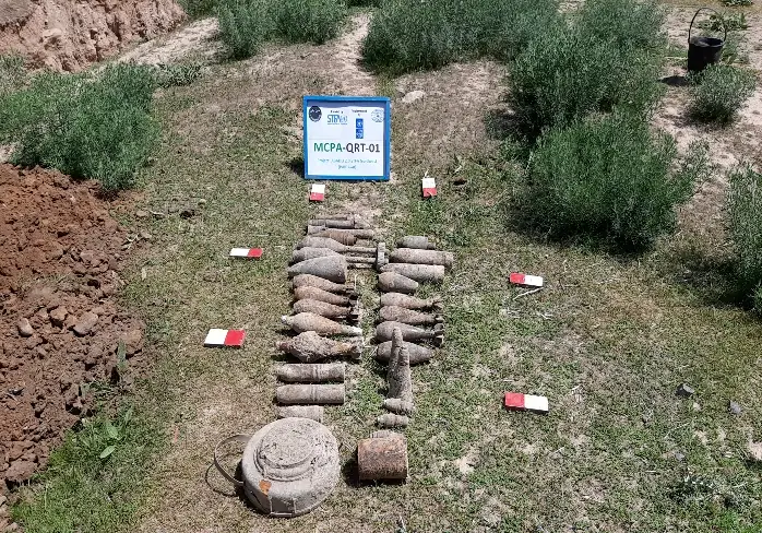 Shells and other explosive items laid out in rows on the ground beside an MCPA team sign, marked with red and white markers.