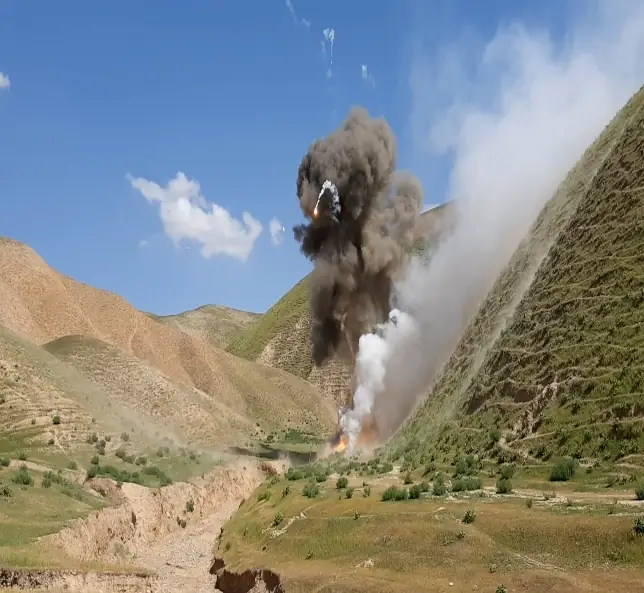 A plume of smoke and dust rises from a controlled explosion in a remote valley.