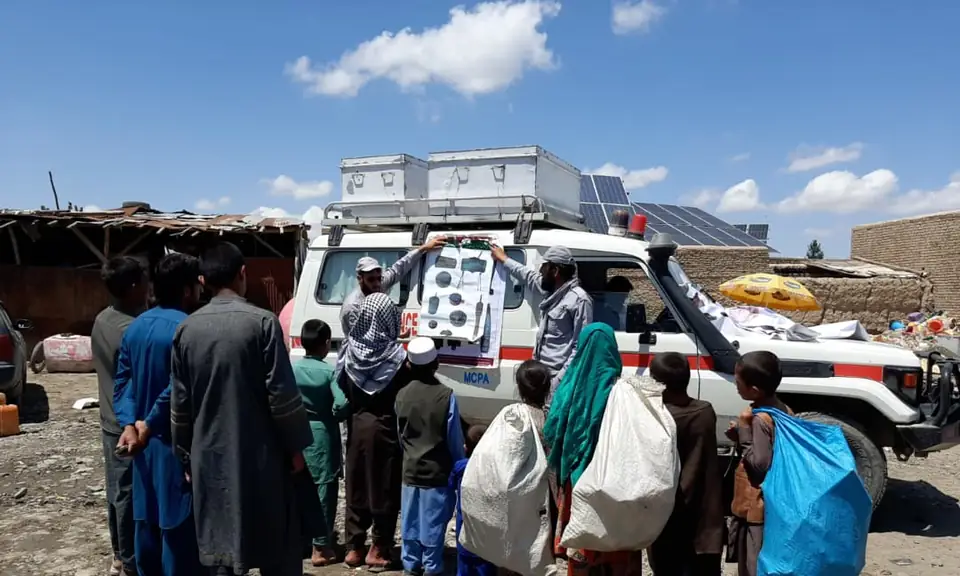 A crowd of men and boys, some carrying sacks of scrap, gather beside an MCPA vehicle displaying a poster of explosive items.