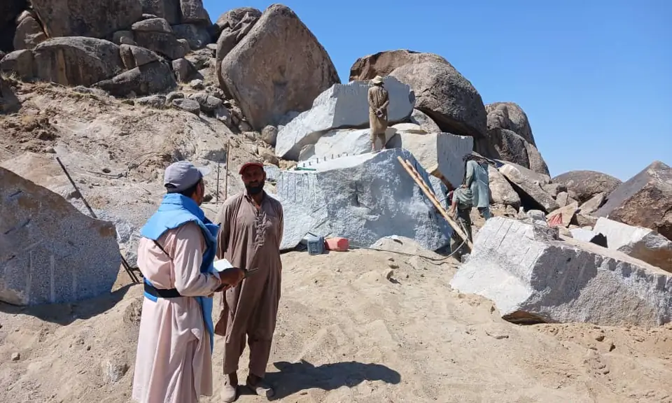 An MCPA deminer in a blue vest talks with a local man among the boulders of the village's stone mine.