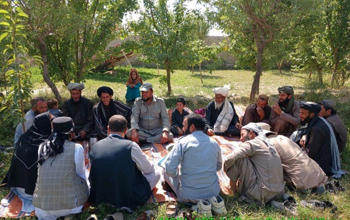 An MCPA quick response team member sitting with village men and elders during a community liaison meeting under trees