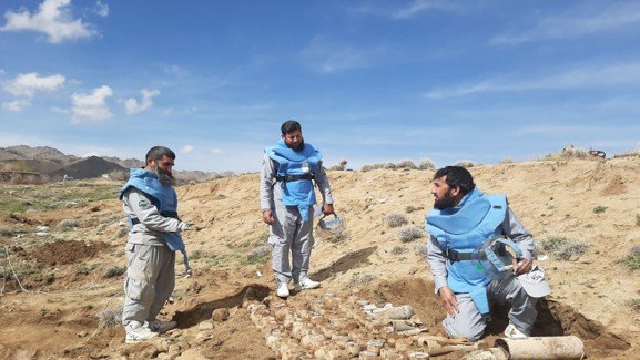 Three MCPA quick response team members in protective vests preparing collected explosive ordnance for destruction on open ground