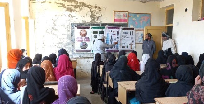 An MCPA educator pointing to a chart of explosive ordnance types for schoolgirls in a classroom