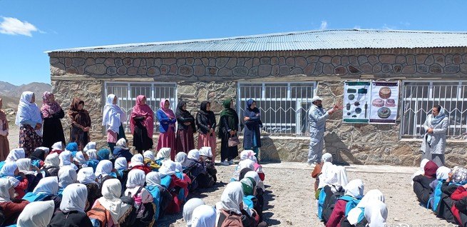Schoolchildren seated in a courtyard while an MCPA educator presents a poster of explosive ordnance types