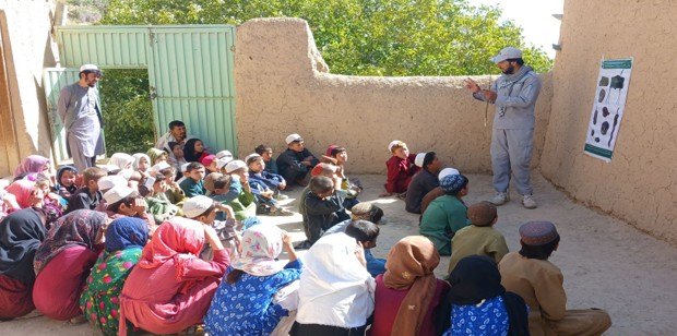 An MCPA educator addressing a mixed group of children seated in a walled school compound