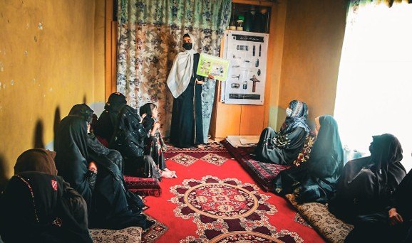 An MCPA risk education session for women seated in a village house, the educator holding up a picture card
